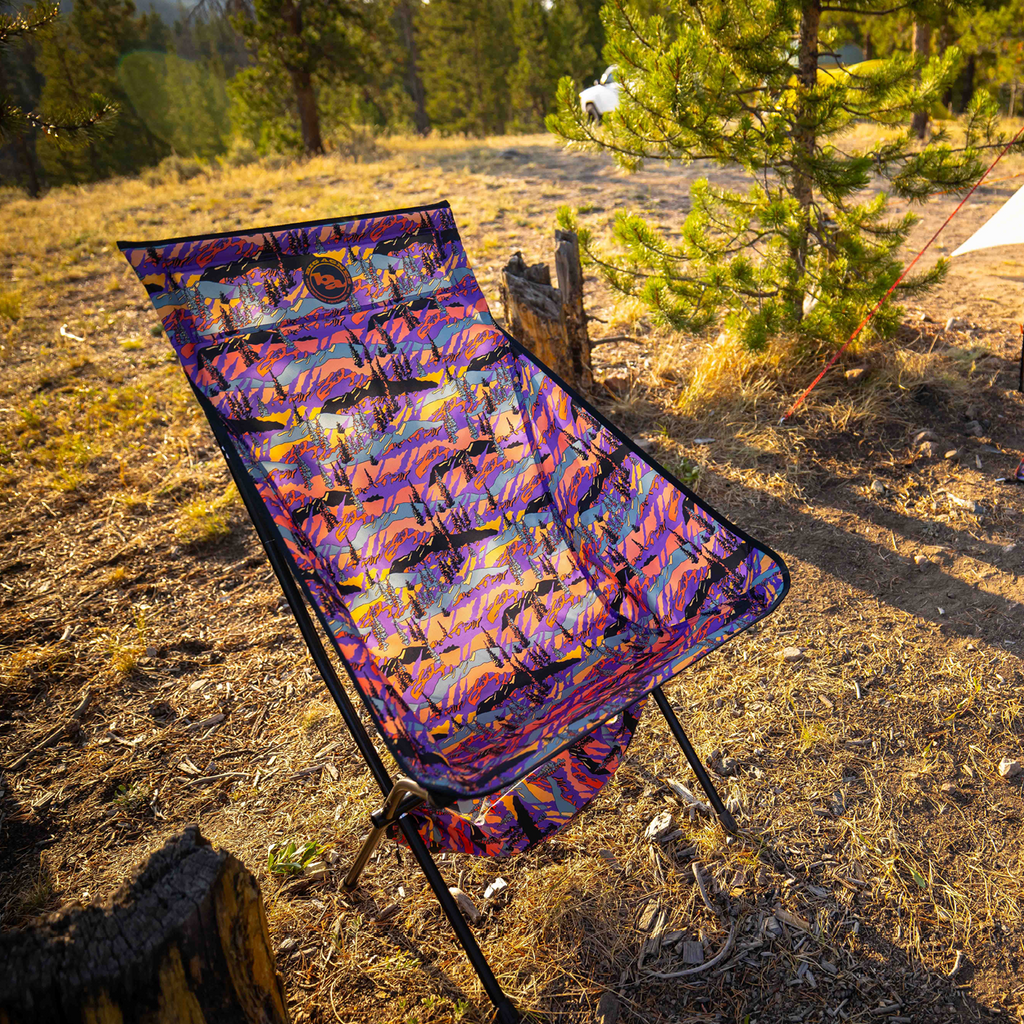 The Big Agnes Big Six Camp Chair - Alpenglow, featuring a colorful, patterned design, is set up on dry grass near pine trees. Sunlight casts warm shadows on the chair and ground, creating an inviting outdoor atmosphere.
