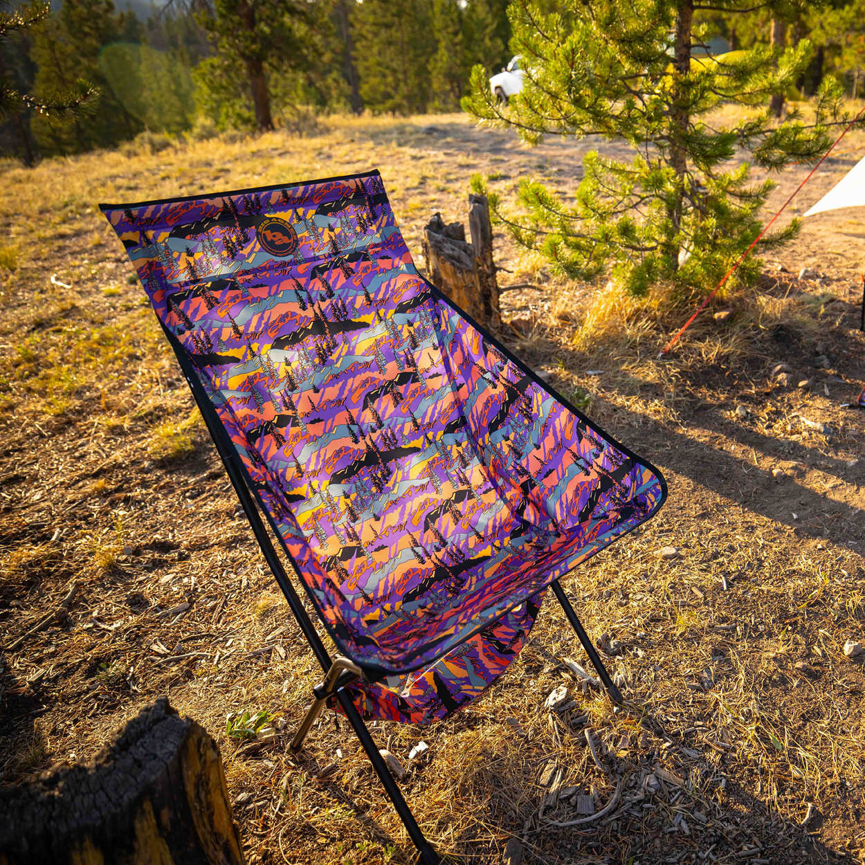 The Big Agnes Returns Big Six Camp Chair - Alpenglow, featuring a colorful, patterned design, is set up on dry grass near pine trees. Sunlight casts warm shadows on the chair and ground, creating an inviting outdoor atmosphere.