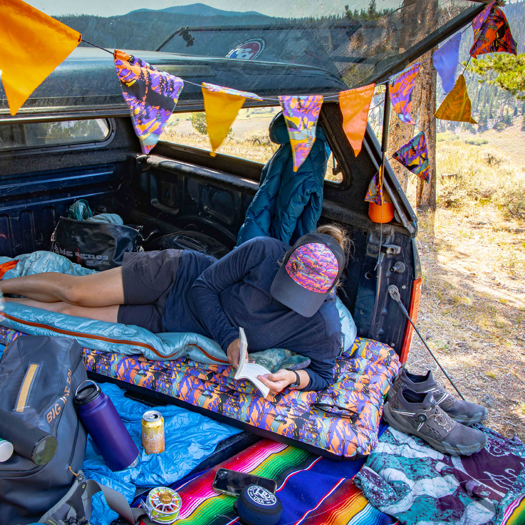 Person lying on a colorful sleeping pad in the back of a vehicle with camping gear and flags.