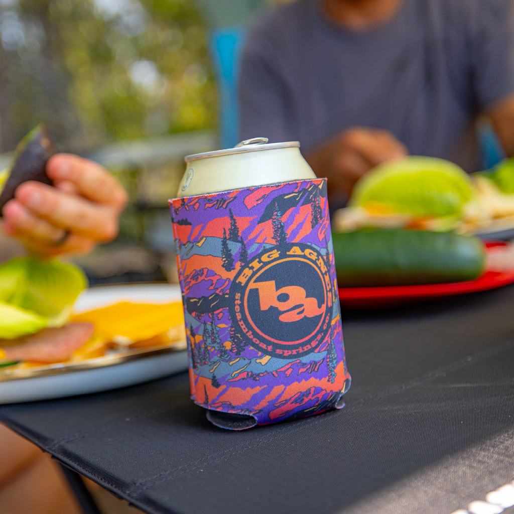 A close-up of a soda can in the limited edition Big Agnes Alpenglow Koozie sits on a picnic table, with plates of food and two people blurred in the background.