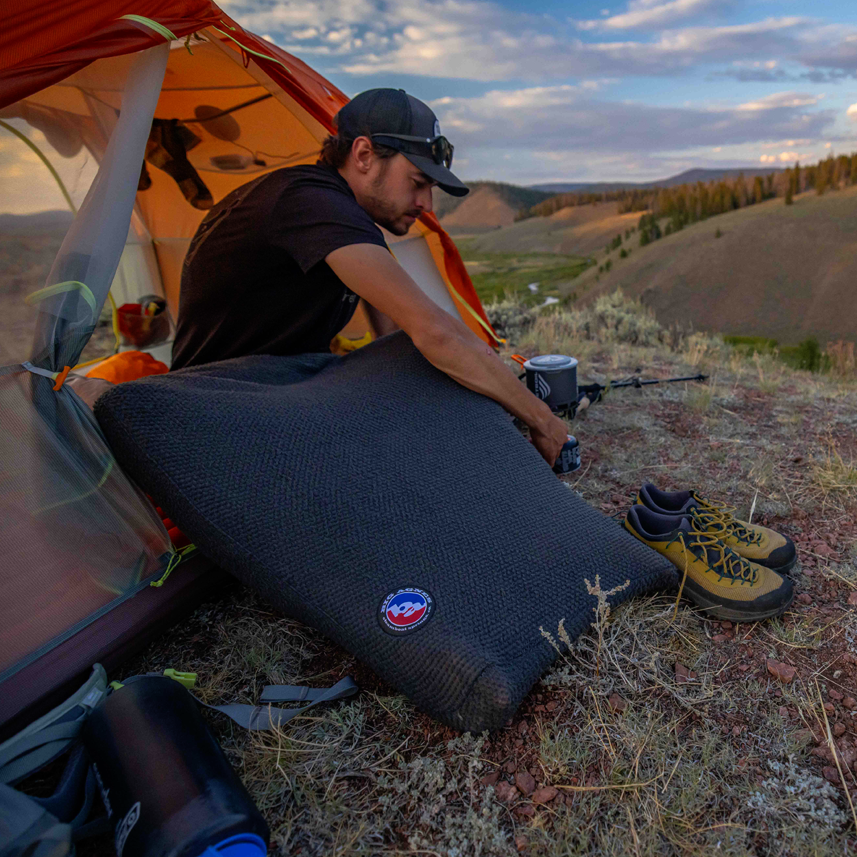 A man sits by an orange tent in hilly terrain, preparing a Big Agnes Alpha Direct Fleece Sleeping Bag Liner. Hiking shoes and water bottles rest on dry grass nearby, with scenic mountains in the background.