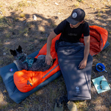 A man in a black shirt and cap sits on a Big Agnes Returns UL Recovery Sleeping Bag Liner laid on the grass, petting a dog beside him. Camping gear and shoes are scattered nearby on the ground.