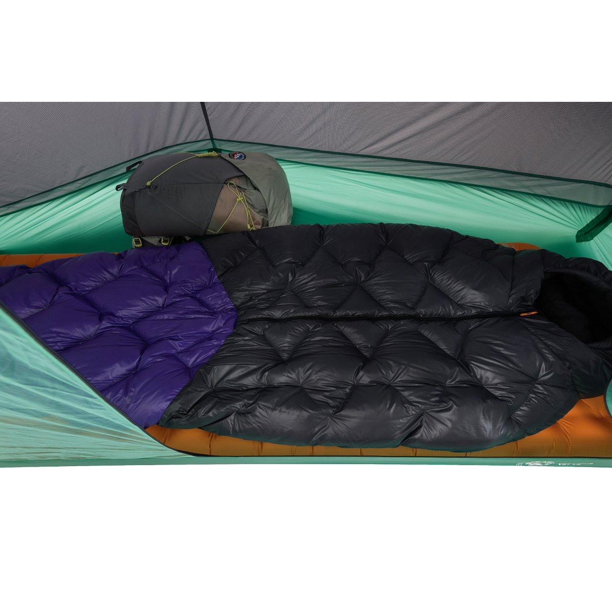 Two sleeping bags—one all black and one with a purple section—are side by side inside a green Big Agnes Pitchpine VST 1.5 tent, set up with an orange sleeping pad beneath and a gray backpack at the head for an ultralight shelter setup.