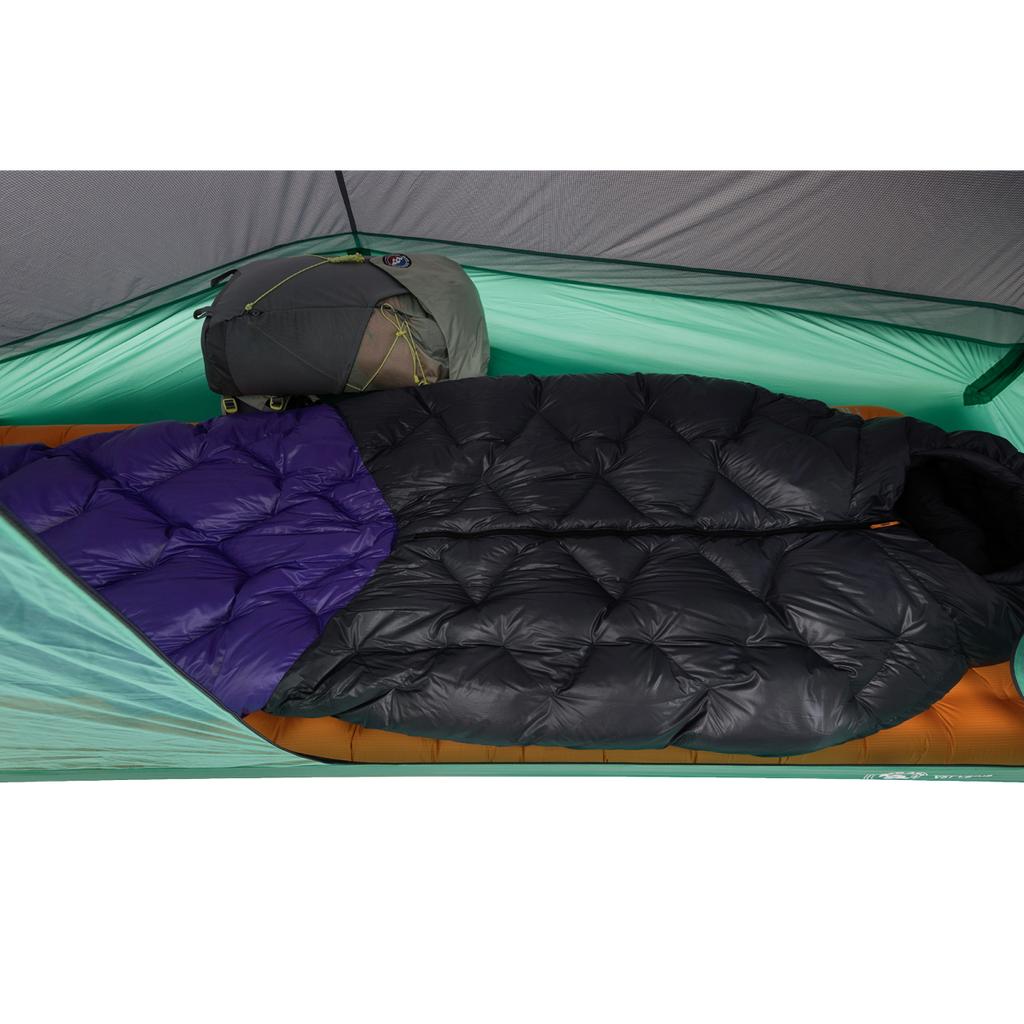 Two sleeping bags—one all black and one with a purple section—are side by side inside a green Big Agnes Returns Pitchpine VST 1.5 tent, set up with an orange sleeping pad beneath and a gray backpack at the head for an ultralight shelter setup.