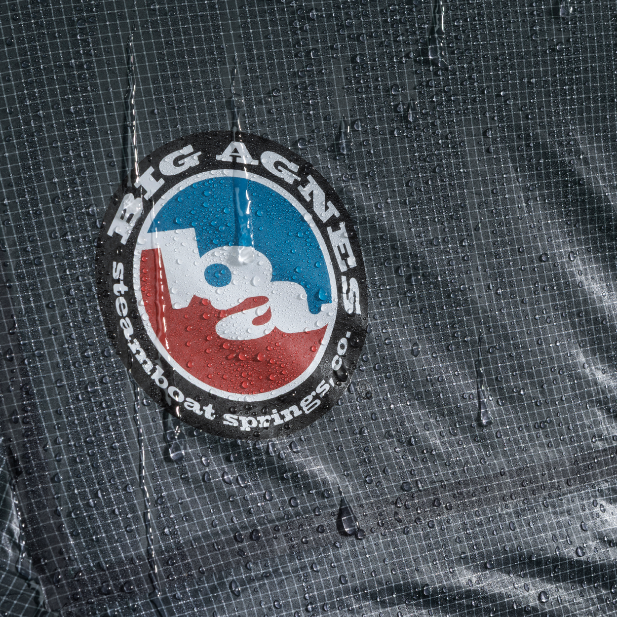 Close-up of wet, grid-patterned Hyperbead fabric with blue and red Big Agnes logo, covered in droplets—ideal for use in the Pitchpine VST 1.5 tent and other ultralight shelters.