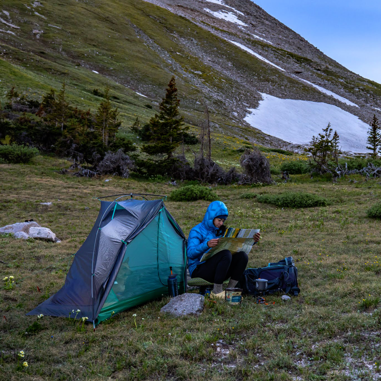 A person in a blue jacket sits beside a green Big Agnes Pitchpine VST 1.5 tent, reading a map on a grassy mountain slope dotted with trees and snow. Ultralight camping gear made with Hyperbead fabric is nearby.