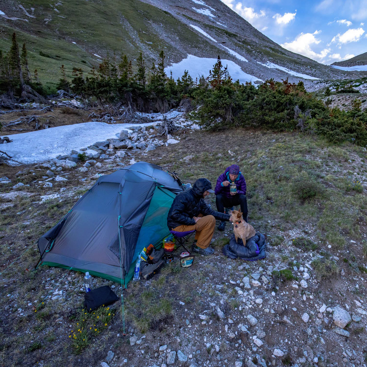 Two people sit beside the green Big Agnes Returns Sarvis VST 2 shelter in snowy mountains, warmly dressed as they pet a small dog on a blanket, surrounded by camping gear and rugged terrain.