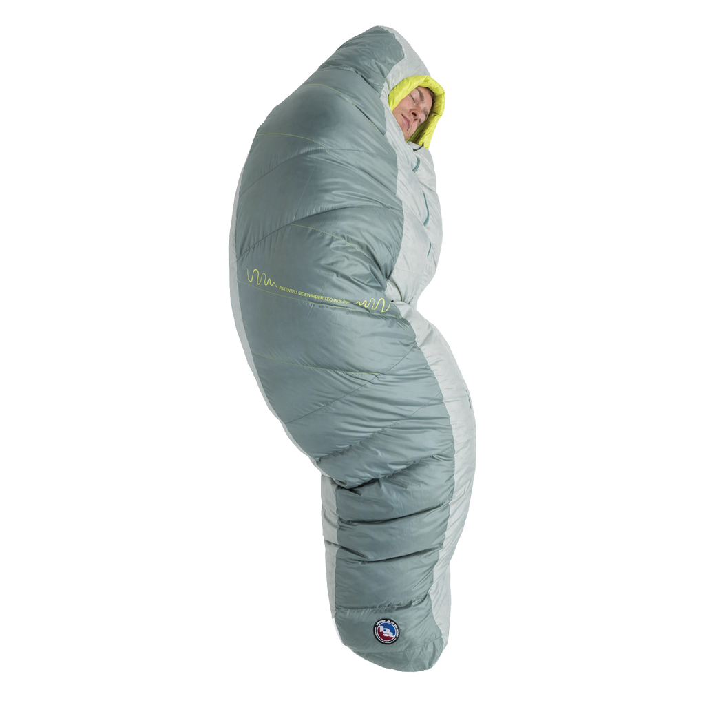 A person stands upright, fully zipped inside the Big Agnes Returns Sidewinder 0˚ sleeping bag in gray and light green, featuring body-mapped insulation with only their face visible.