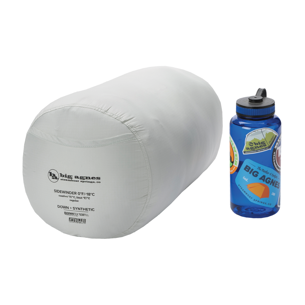 A light gray Big Agnes Returns Sidewinder 0˚ sleeping bag with body-mapped insulation, packed in a cylindrical stuff sack, sits next to a blue sticker-covered Nalgene bottle on a white background.