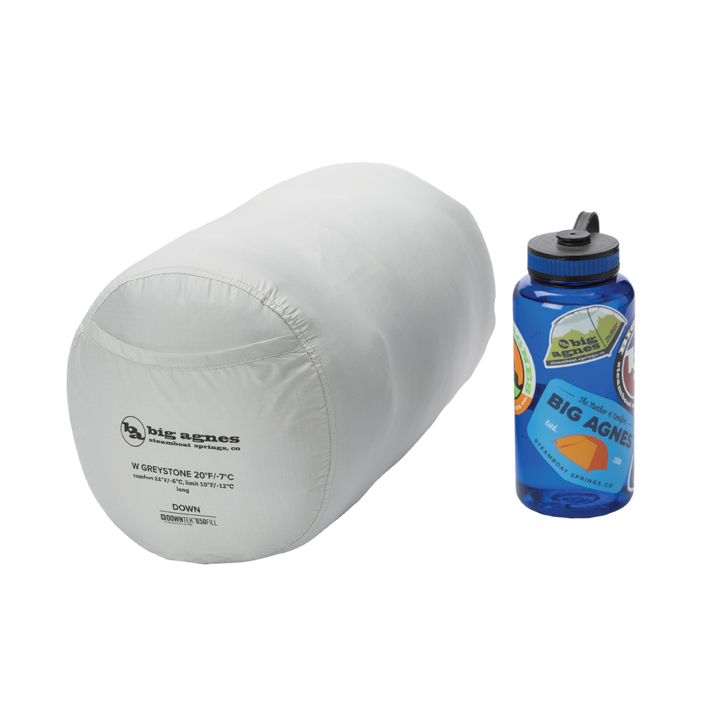 A packed Big Agnes Women's Greystone 30° sleeping bag is shown next to a blue Nalgene water bottle for size comparison, both placed on a plain white background.