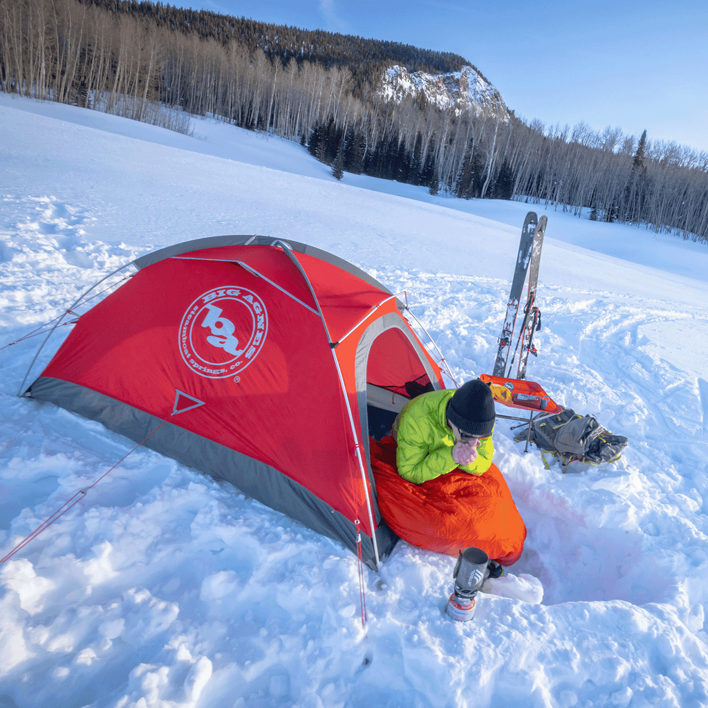 A person sits in a bright orange sleeping bag beside the red Big Agnes Returns Shield 2 tent on snow, sipping from a mug. Skis, poles, and gear are nearby with bare trees, a snowy landscape, and a mountain in the background.