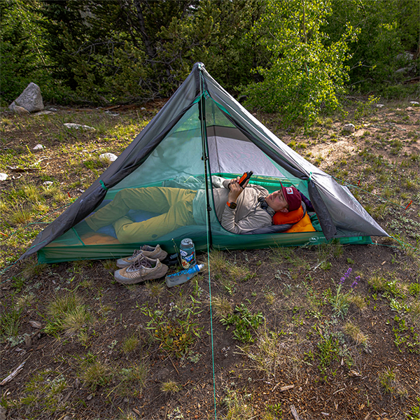 Inside a Big Agnes Returns String Ridge VST 1.5 tent, a person relaxes on a sleeping pad with a book. Nearby, hiking shoes and a water bottle sit outside the tent on uneven grass, surrounded by green trees and bushes.