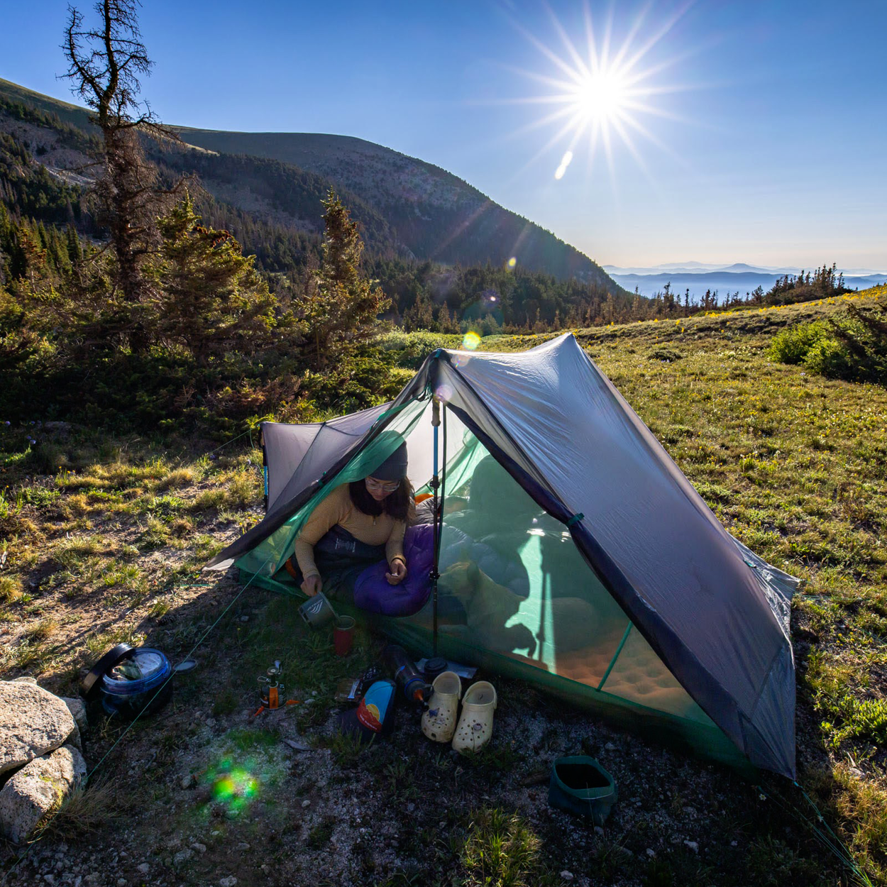 A person relaxes inside a Big Agnes Returns String Ridge VST 2.5 ultralight tent set on grassy mountain terrain, surrounded by camping gear and pine trees, with distant hills under a sunny, clear sky.